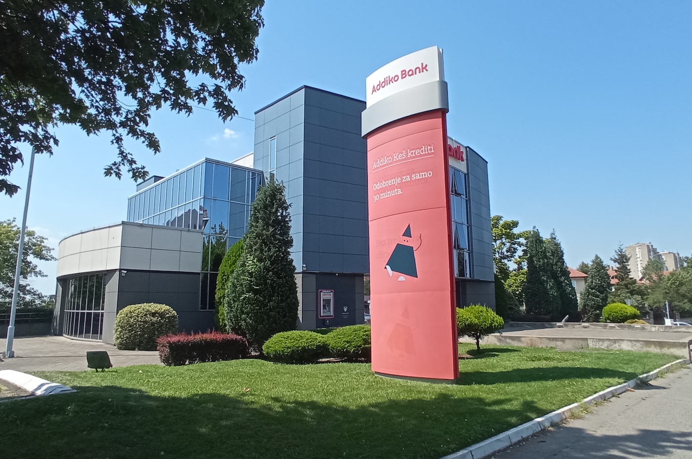 Photo of Addiko Bank - a large glass building with a tall red sign in front and lush green landscaping.