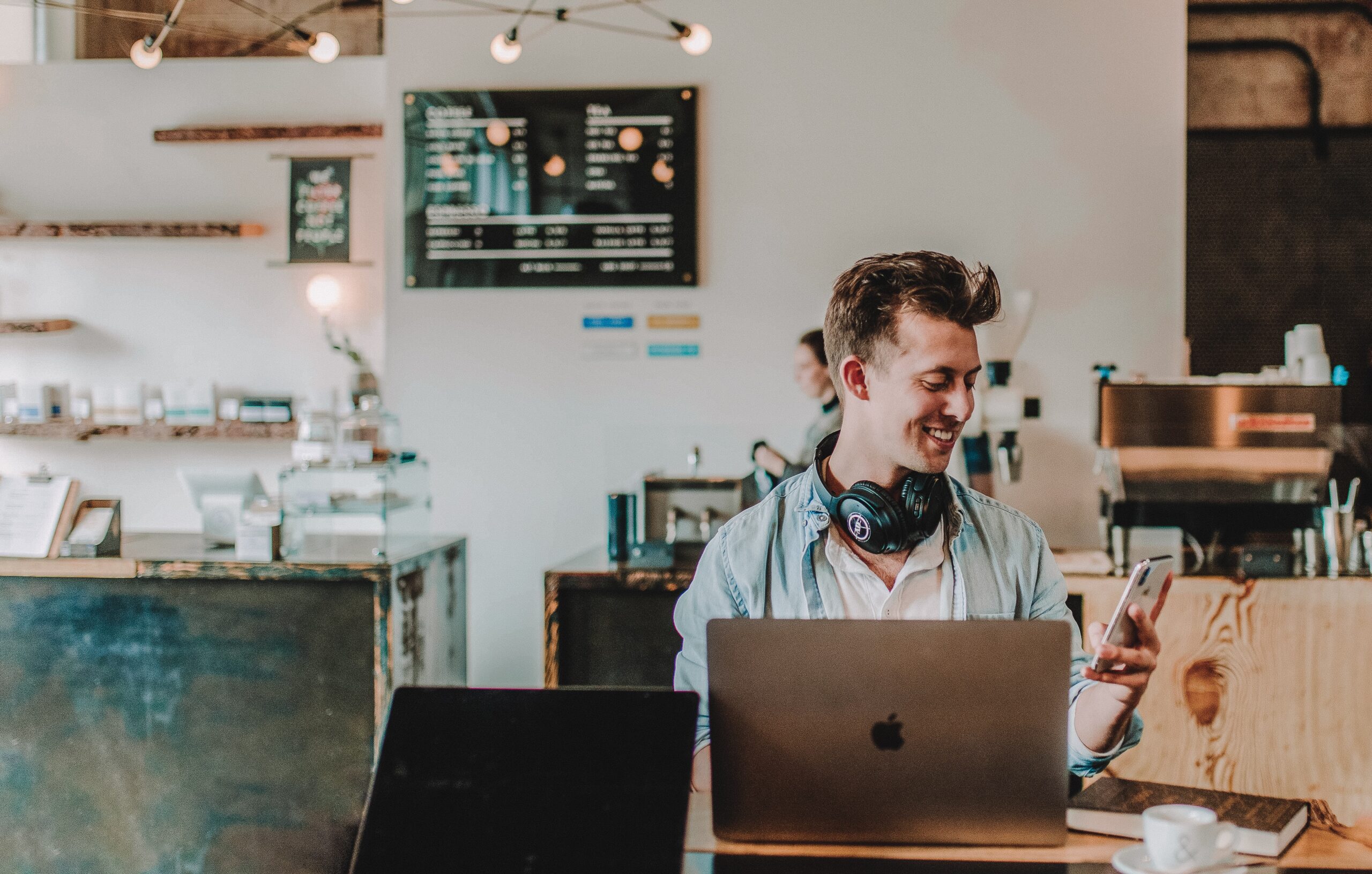 Photo of a man in a coffee shop with a laptop looking at a mobile phone